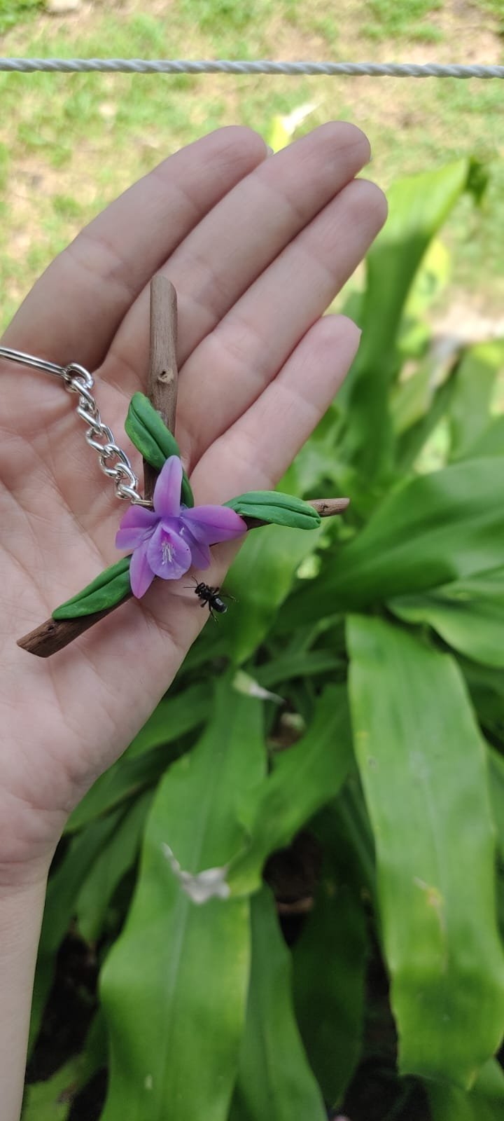 Hand holding a small purple flower with green sepals and a wooden clothespin charm, with a dark insect on it, in a garden setting