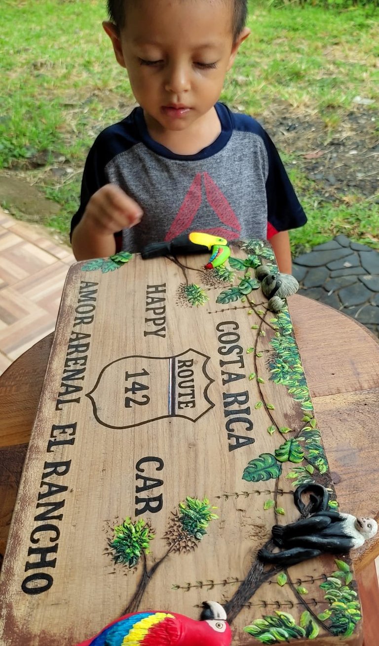 Young child exploring a wooden craft plaque with text and painted green plants, holding a small toy tool outdoors