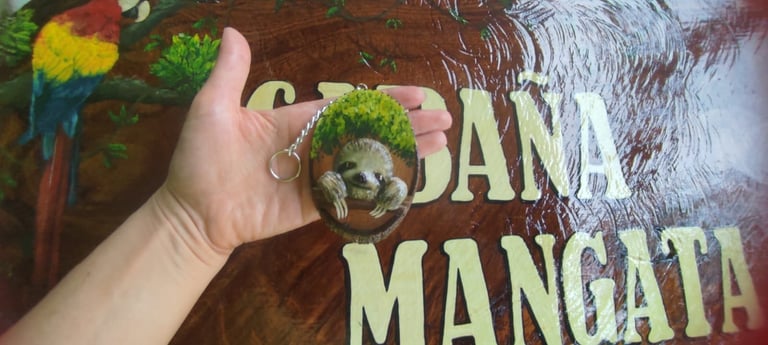 Hand holding a small sloth figurine in front of a Rana Mangata text and jungle-themed mural with parrots