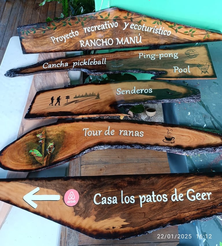 Stack of wooden directional signs with Spanish text pointing to local attractions and activities like Rancho Manu, ping-pong, and Casa los patos de Geer