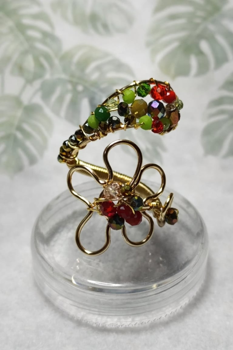 Two gold wire brooches with colorful beads displayed on a clear acrylic stand against a leaf-patterned background