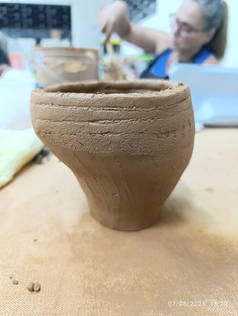 Terracotta clay vessel with textured surface on table, person in background working with pottery