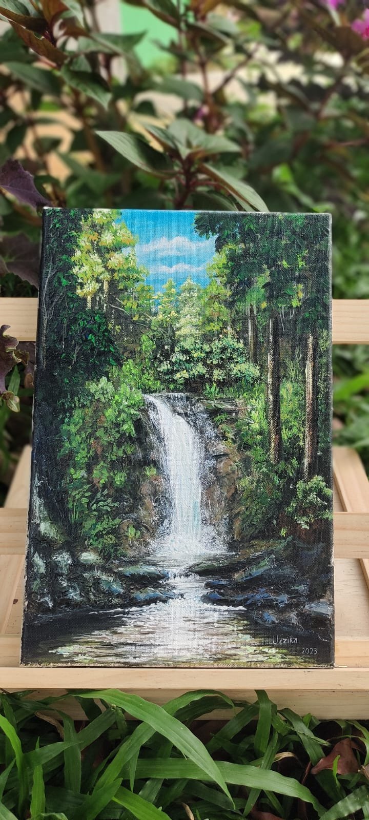 Framed oil painting of a forest waterfall surrounded by lush green trees displayed outdoors on a wooden shelf among potted plants