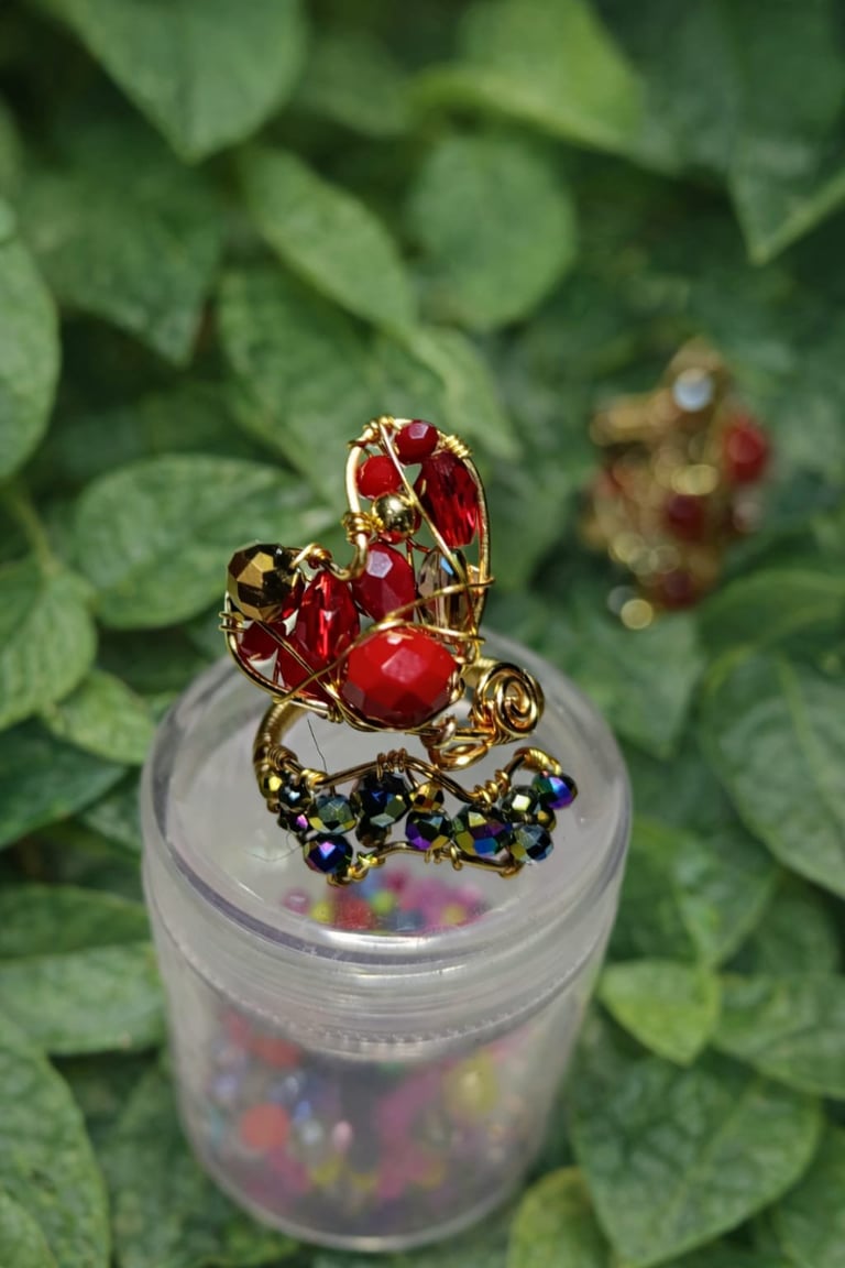 Gold ring with red gemstones and blue crystals displayed on clear container, green foliage background