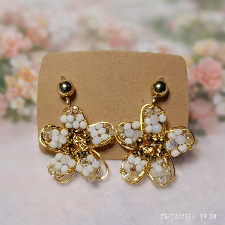 Pair of gold flower-shaped earrings with white beads and crystals on a tan display card, photographed on a floral background