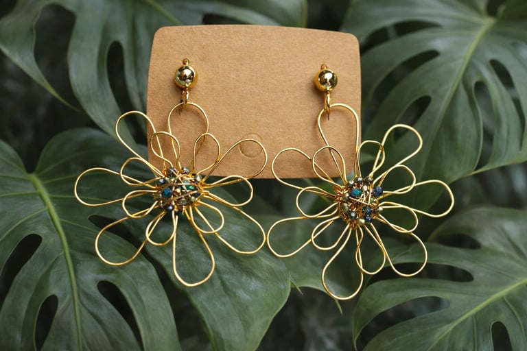 Gold wire daisy flower earrings with blue gemstone centers displayed on tan card against green monstera leaves